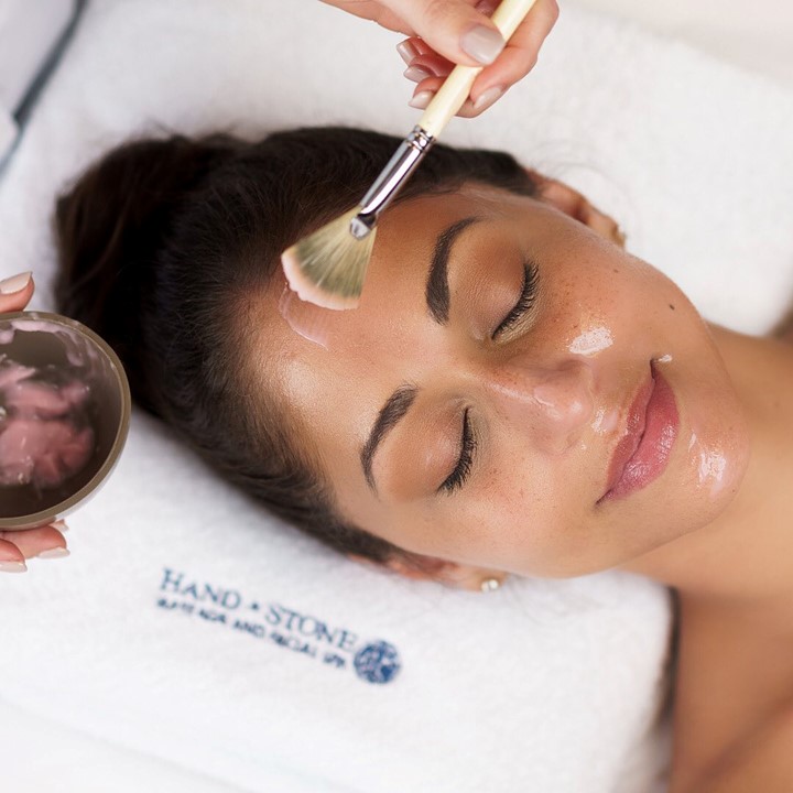 A woman rests her head on a towel as an esthetician spreads a skin treatment on her face with a fan brush.